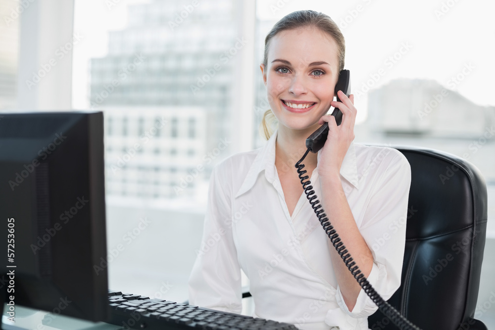 Smiling businesswoman sitting at desk on the phone looking at ca Smiling businesswoman sitting at desk on the phone looking at ca