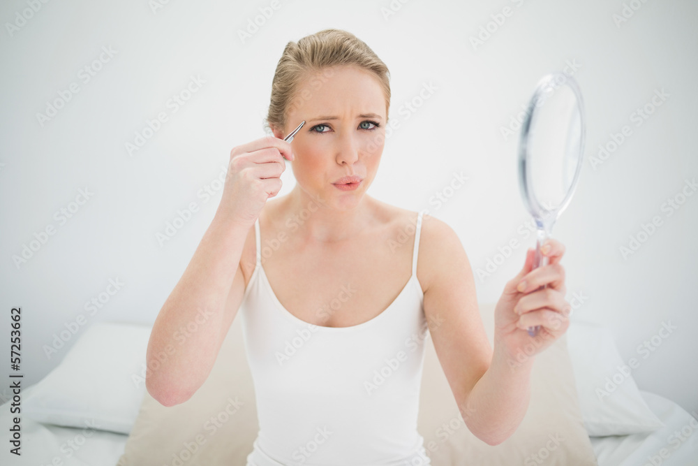 Natural frowning blonde holding mirror and using tweezers Natural frowning blonde holding mirror and using tweezers