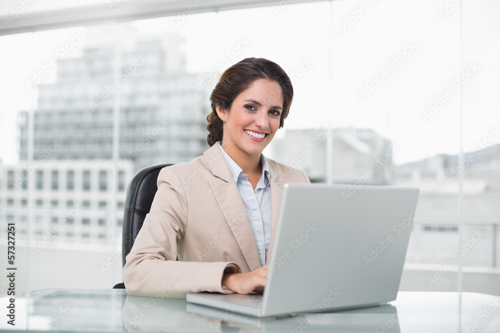 Happy businesswoman typing on laptop at her desk looking at came Happy businesswoman typing on laptop at her desk looking at came