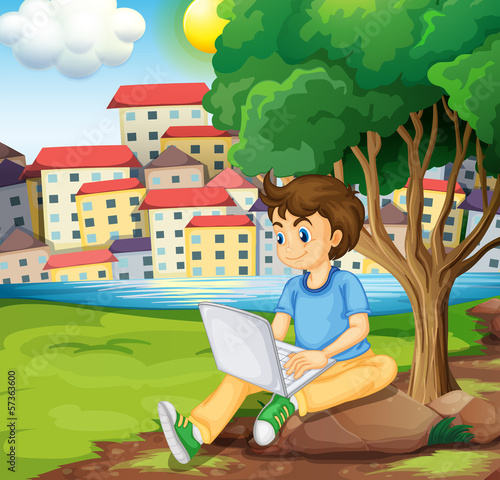 A young boy using the laptop under the tree at the riverbank