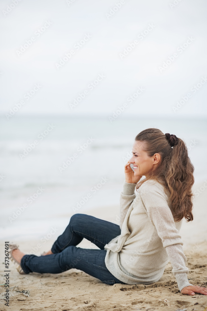 Happy young woman sitting on lonely beach and talking cell phone