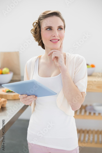 Smiling thinking woman holding her tablet standing in kitchen