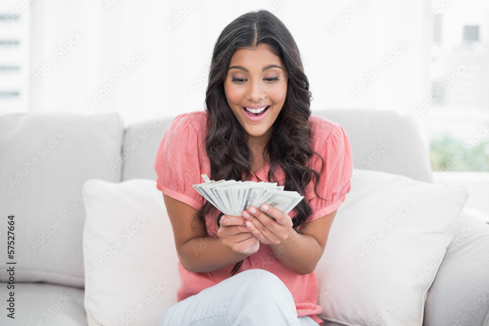 Excited cute brunette sitting on couch holding money Excited cute brunette sitting on couch holding money
