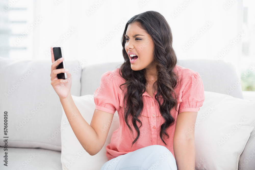 Angry cute brunette sitting on couch holding smartphone Angry cute brunette sitting on couch holding smartphone