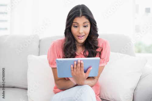 Excited cute brunette sitting on couch using tablet