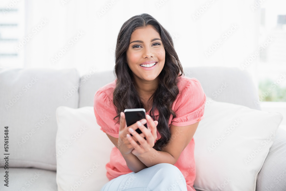 Cheerful cute brunette sitting on couch holding smartphone Cheerful cute brunette sitting on couch holding smartphone