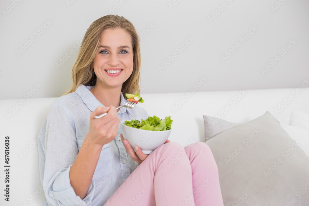 Smiling casual blonde sitting on couch holding salad bowl Smiling casual blonde sitting on couch holding salad bowl