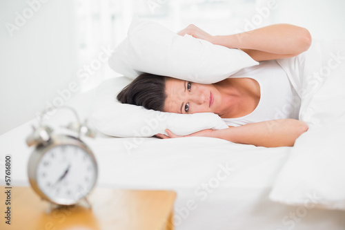 Woman covering ears with pillow and alarm clock on side table