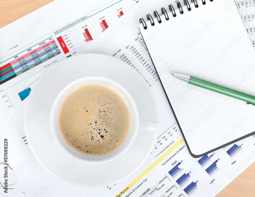 Coffee cup and notepad over papers with numbers and charts Coffee cup and notepad over papers with numbers and charts