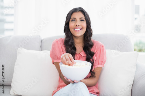 Cheerful cute brunette sitting on couch holding popcorn bowl