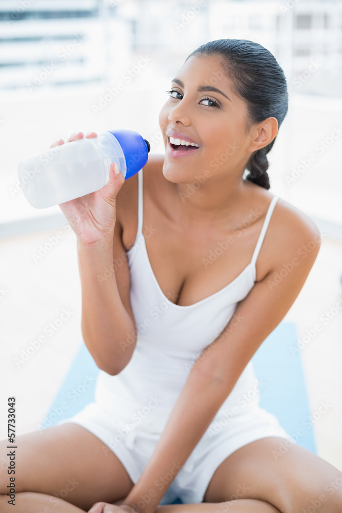 Smiling toned brunette sitting on floor holding sports bottle Smiling toned brunette sitting on floor holding sports bottle