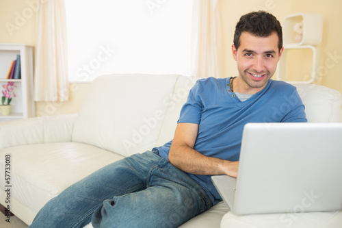Casual smiling man sitting on couch using laptop looking at came