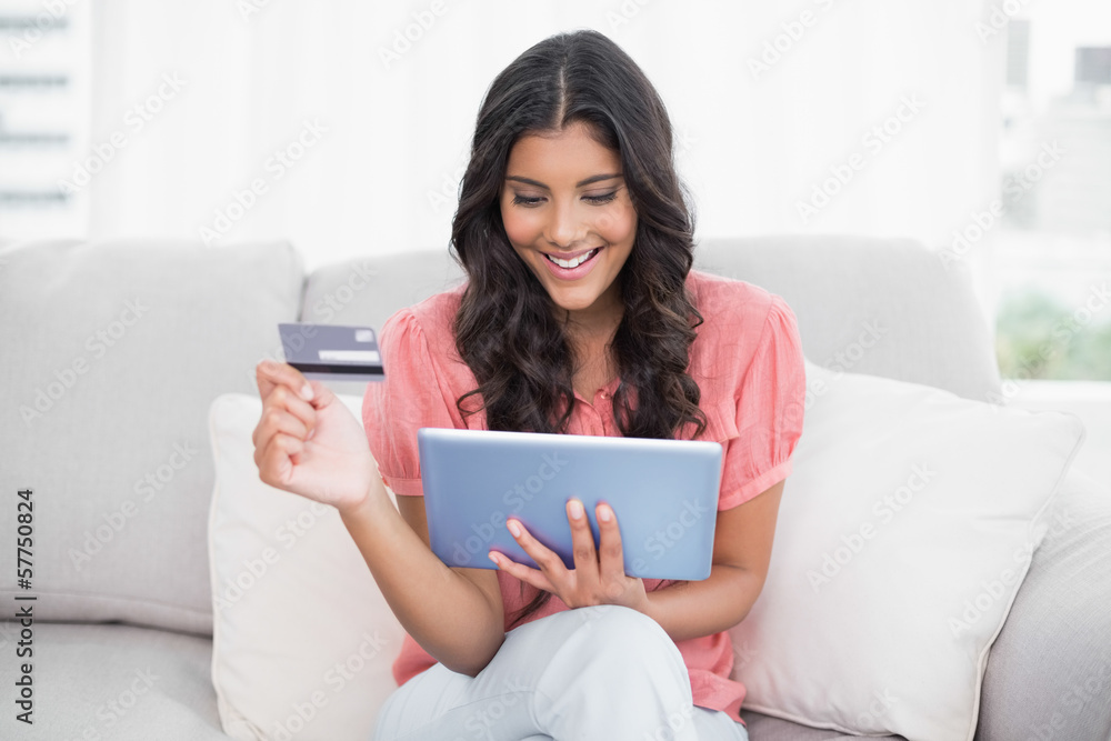 Smiling cute brunette sitting on couch holding credit card and t Smiling cute brunette sitting on couch holding credit card and t