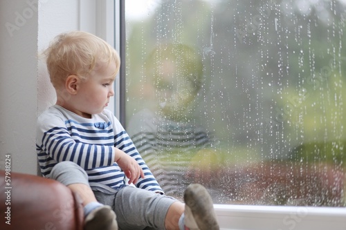 Cute baby girl looking outside through the window during rain