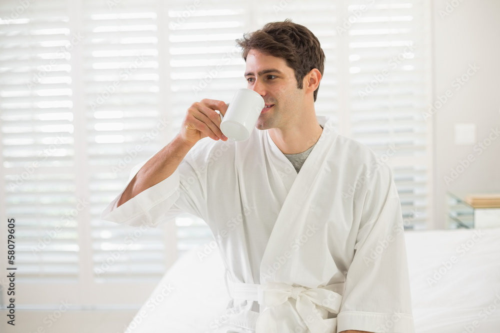 Young man in bathrobe drinking coffee in bedroom