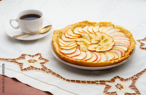 A tasty golden homemade apple tart and a cup of black coffee on an embroidered tablecloth