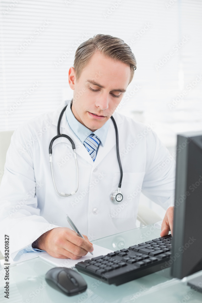 Doctor writing a note while using computer at medical office Doctor writing a note while using computer at medical office