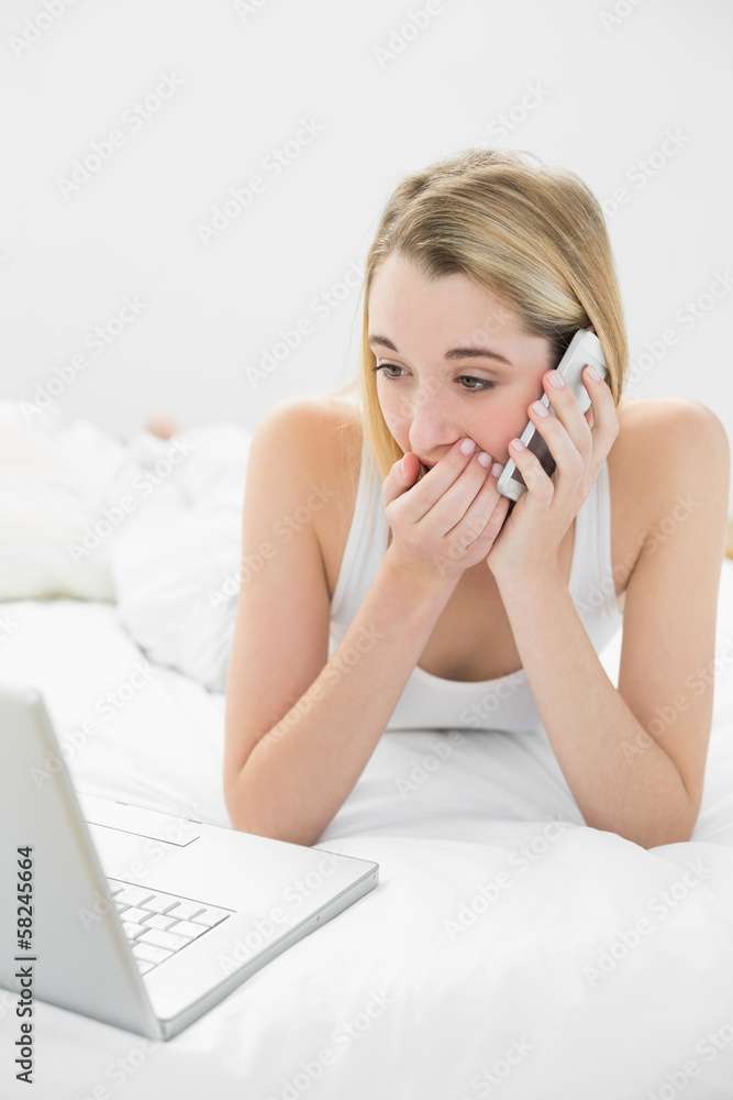 Blonde shocked woman phoning and using her notebook lying on her Blonde shocked woman phoning and using her notebook lying on her