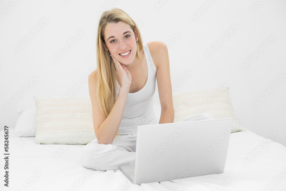Content blonde woman using her notebook sitting on her bed smili Content blonde woman using her notebook sitting on her bed smili