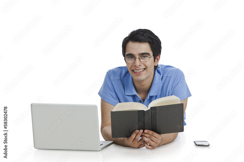 Happy man lying in front of a laptop and reading a book Happy man lying in front of a laptop and reading a book