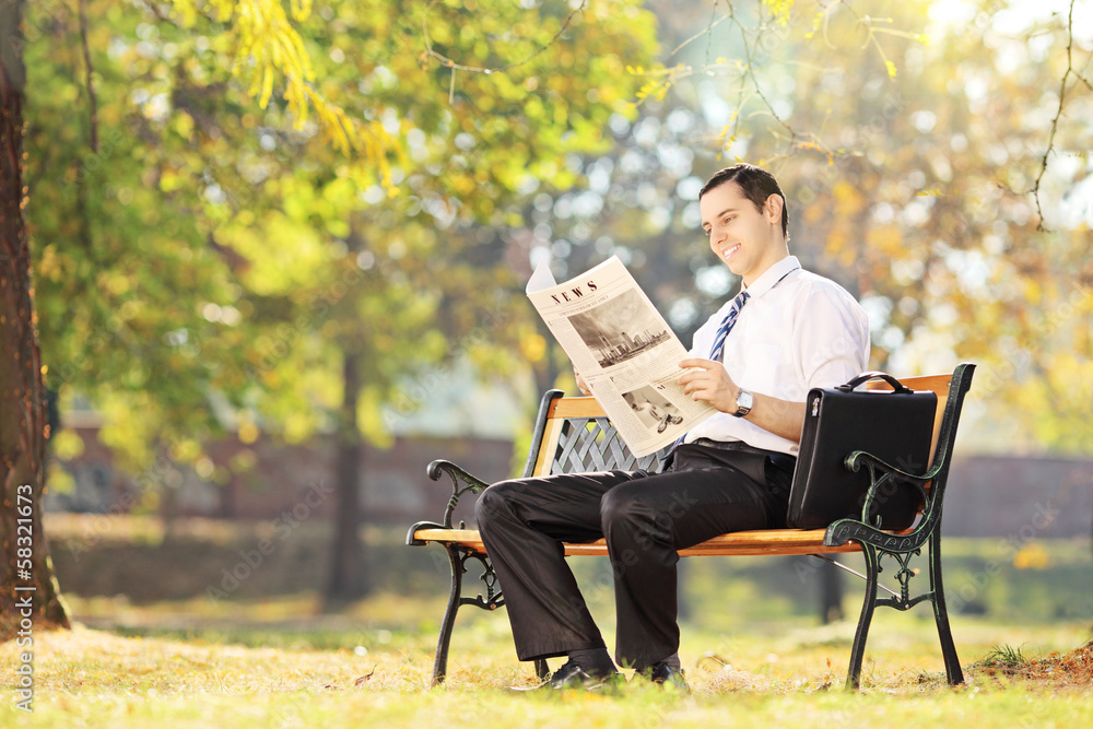 Young smiling man seated on bench reading newspaper in a park Young smiling man seated on bench reading newspaper in a park