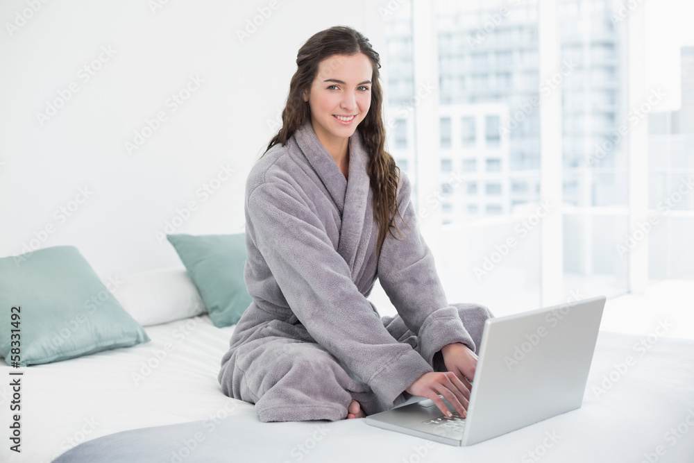 Smiling brunette in bathrobe using laptop in bed Smiling brunette in bathrobe using laptop in bed