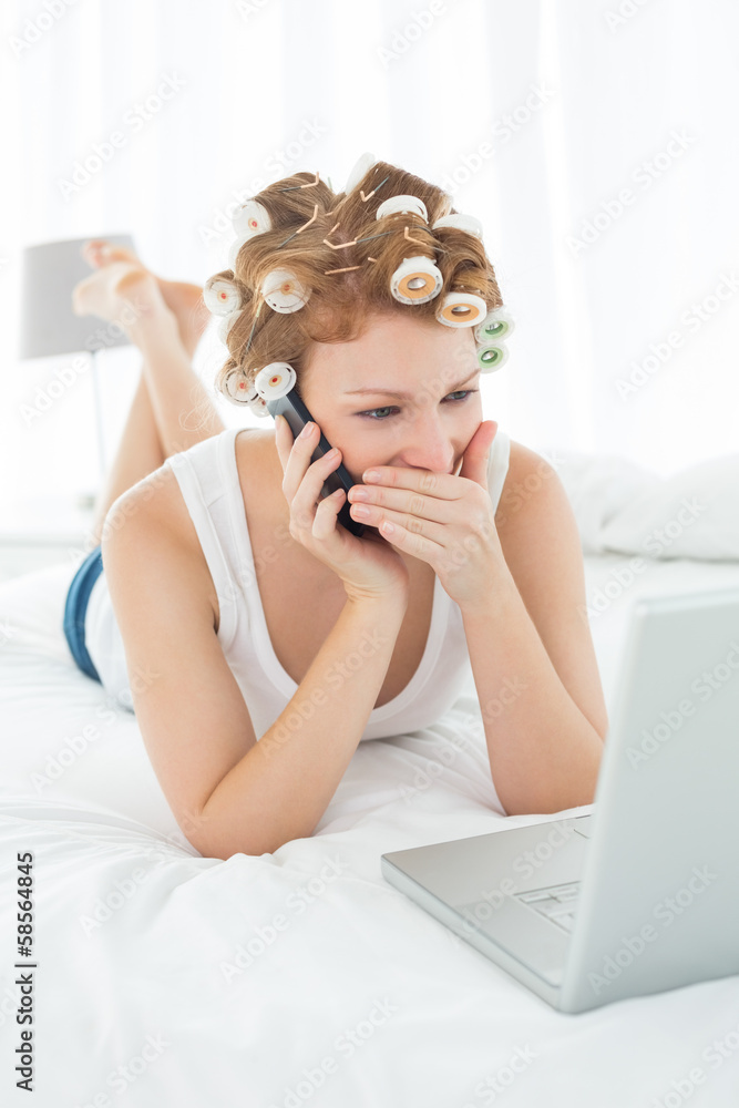 Shocked woman in hair curlers using cellphone and laptop in bed