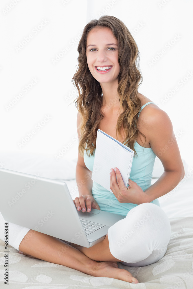 Naklejka premium Woman using laptop as she holds a notepad in bed