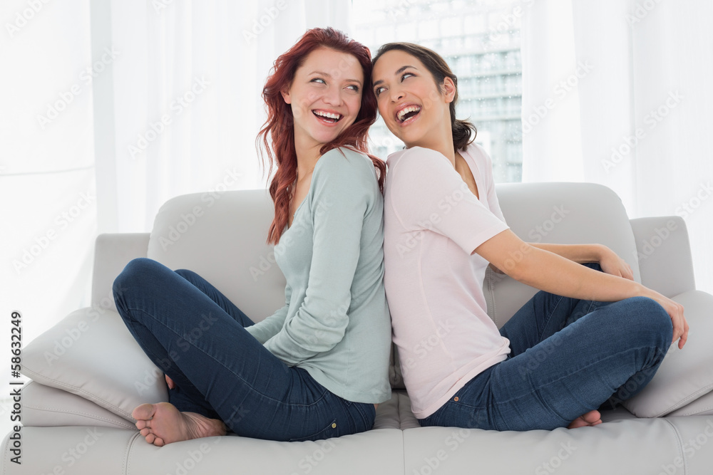Cheerful young female friends sitting back to back in the living