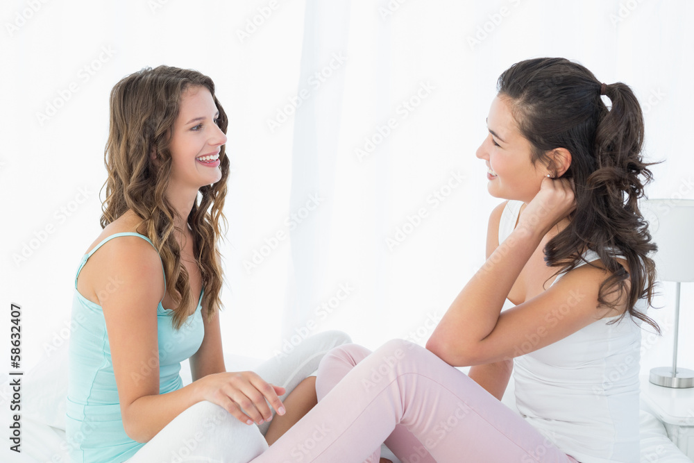 Happy young female friends sitting chatting on bed