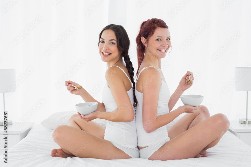 Two smiling female friends with bowls sitting on bed Two smiling female friends with bowls sitting on bed