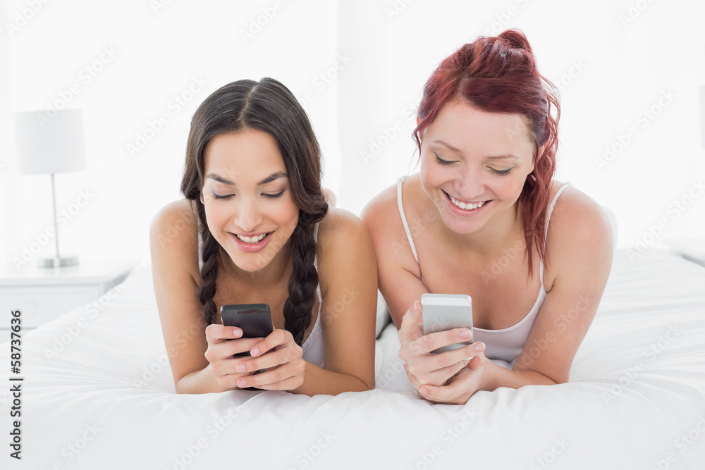 Smiling female friends text messaging on bed Smiling female friends text messaging on bed