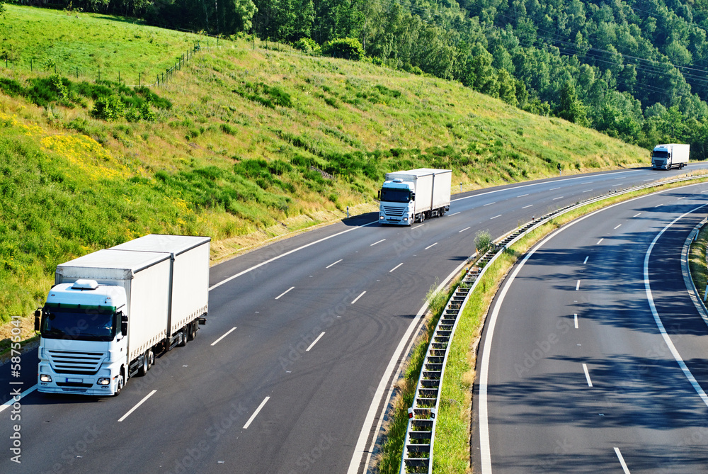 Highway with three oncoming white truck in a wooded landscape