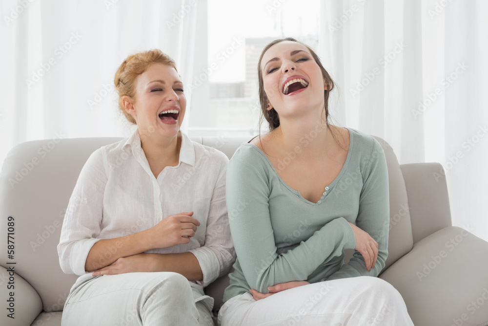 Female friends laughing while sitting on sofa in the living room Female friends laughing while sitting on sofa in the living room