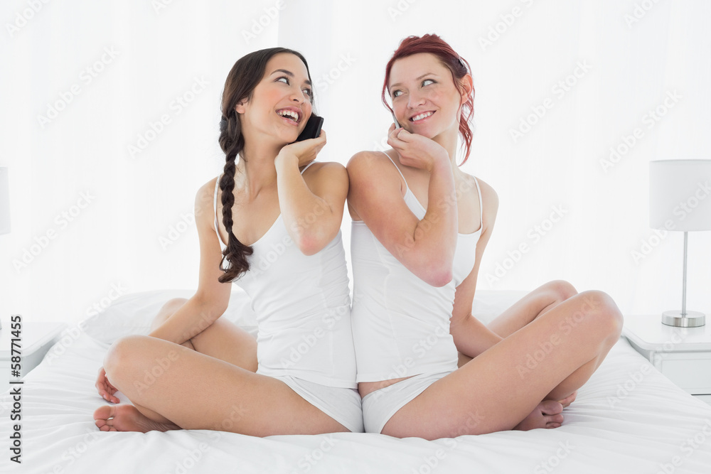 Friends using mobile phone while sitting back to back on bed Friends using mobile phone while sitting back to back on bed