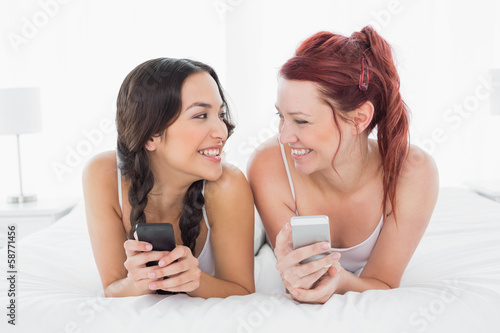 Two smiling female friends text messaging on bed