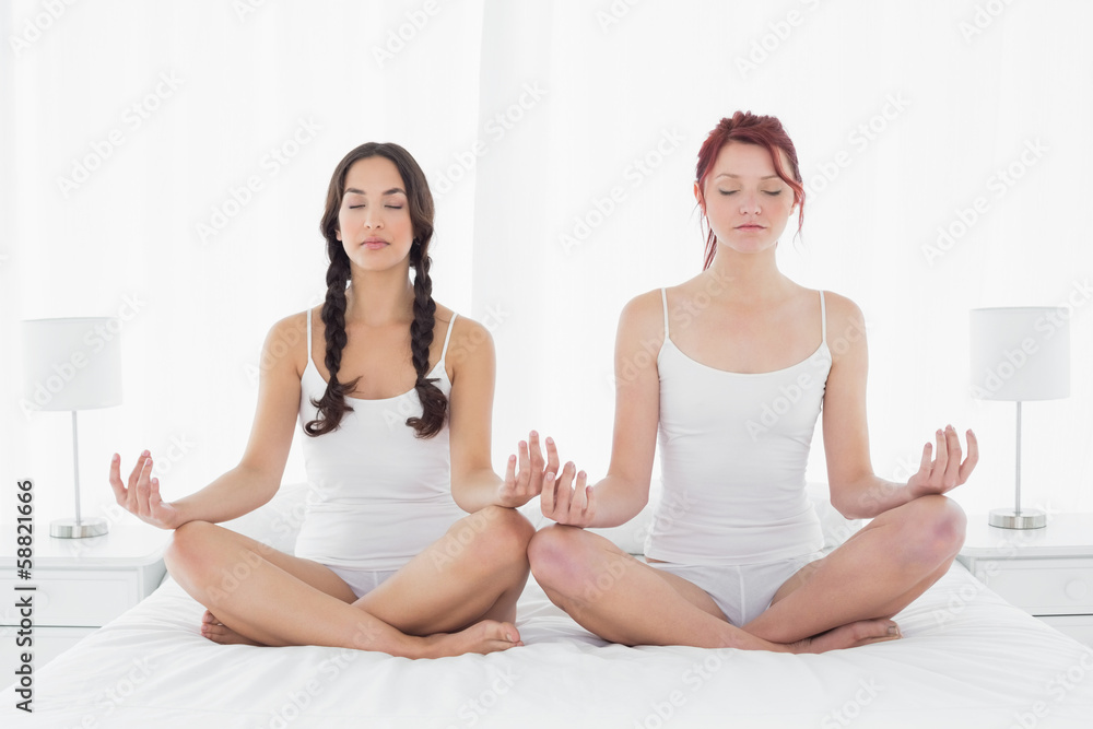 Two women with eyes closed sitting in lotus posture on bed Two women with eyes closed sitting in lotus posture on bed