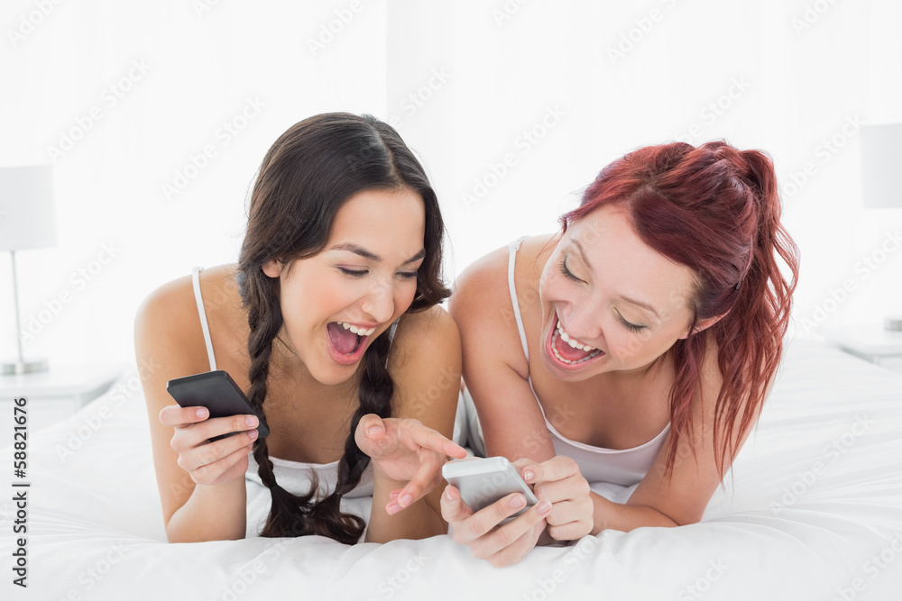 Cheerful young female friends text messaging on bed Cheerful young female friends text messaging on bed