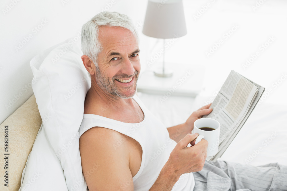 Smiling mature man with coffee cup and newspaper in bed Smiling mature man with coffee cup and newspaper in bed