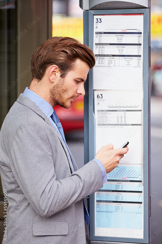Businessman At Bus Stop With Mobile Phone Reading Timetable Businessman At Bus Stop With Mobile Phone Reading Timetable
