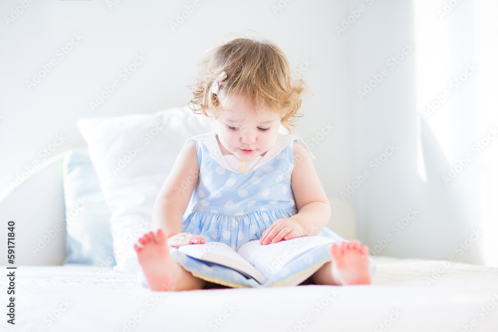 Funny toddler girl in a blue dress reading a book on a white bed Funny toddler girl in a blue dress reading a book on a white bed