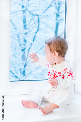 Baby in a sweater with Christmas ornament sitting at a window