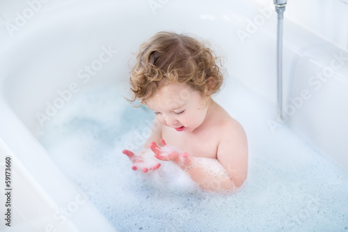 Little baby girl with curly hair playing with foam in a bath