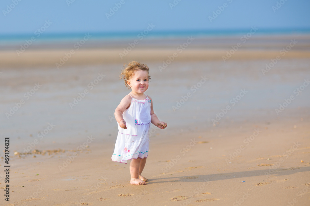 Curly baby girl wearing a white festive dress walking on a beach Curly baby girl wearing a white festive dress walking on a beach