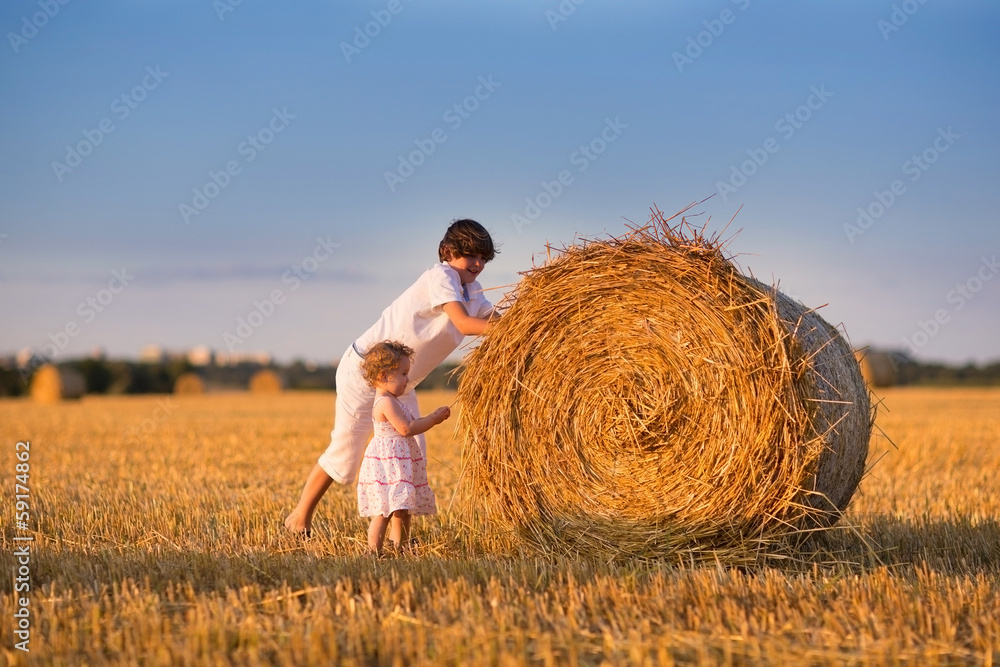 Brother and baby sister pushing hay bales in a field at sunset Brother and baby sister pushing hay bales in a field at sunset
