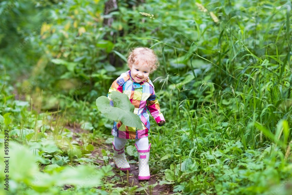 Cute curlu baby girl playing with big leaves in a park on a rain