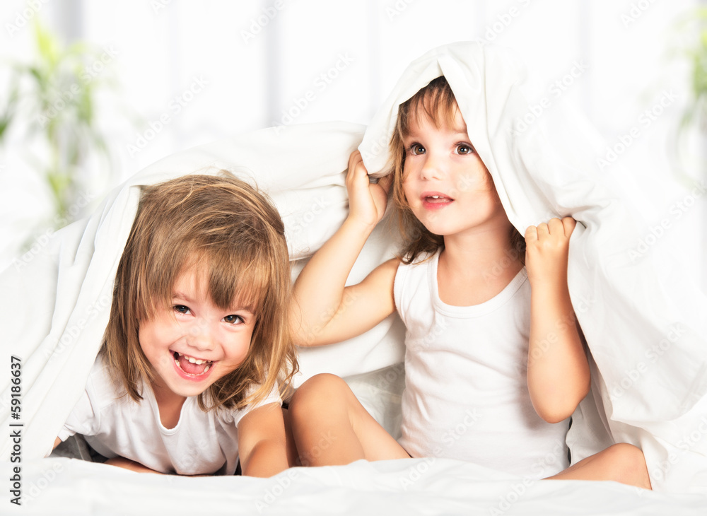 happy little girls twins sister in bed under the blanket having happy little girls twins sister in bed under the blanket having