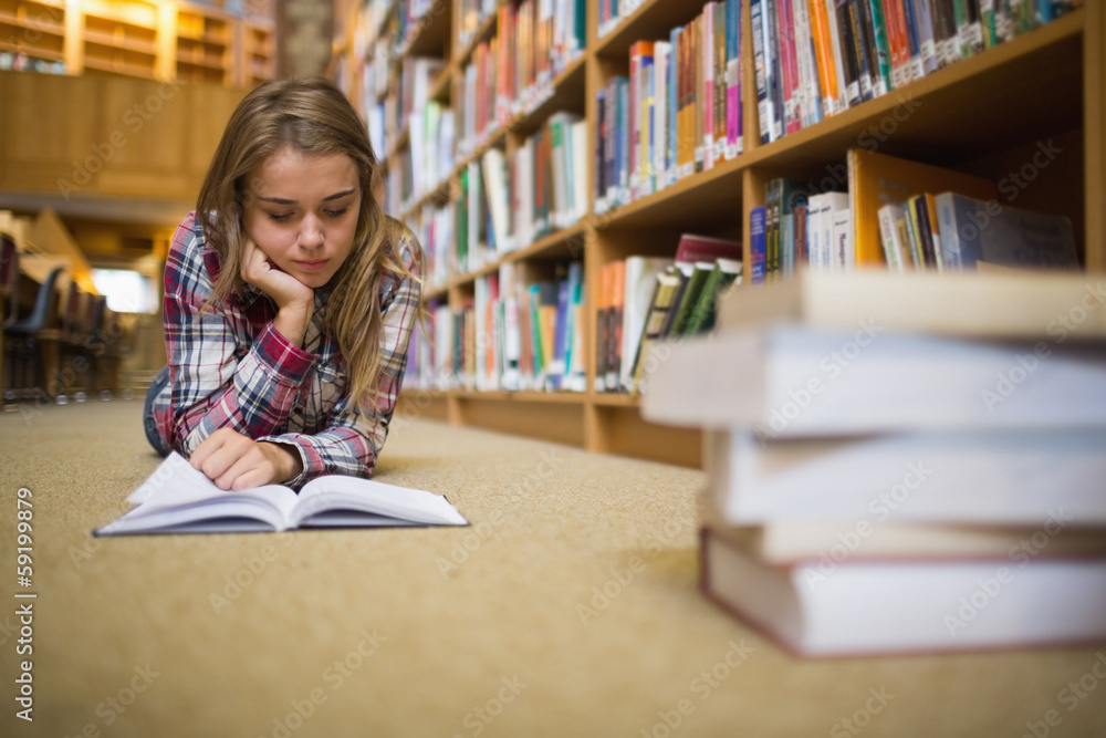 Pretty focused student lying on library floor reading book Pretty focused student lying on library floor reading book