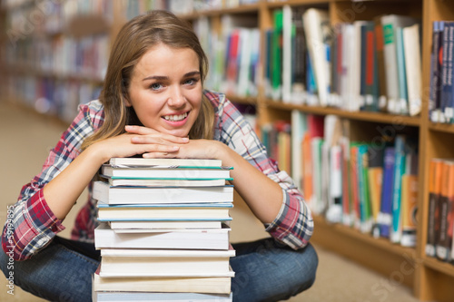 Smiling student sitting on library floor leaning on pile of book