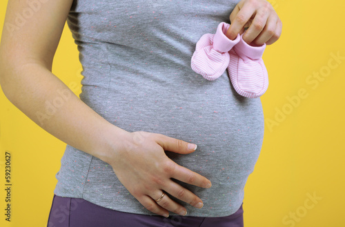 Pregnant woman holding pink baby shoes on yellow background